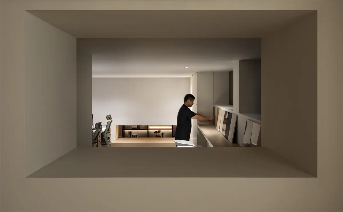 View through a rectangular opening into a serene minimalist home office featuring clean lines, hidden storage, warm LED lighting, and a man arranging artwork.