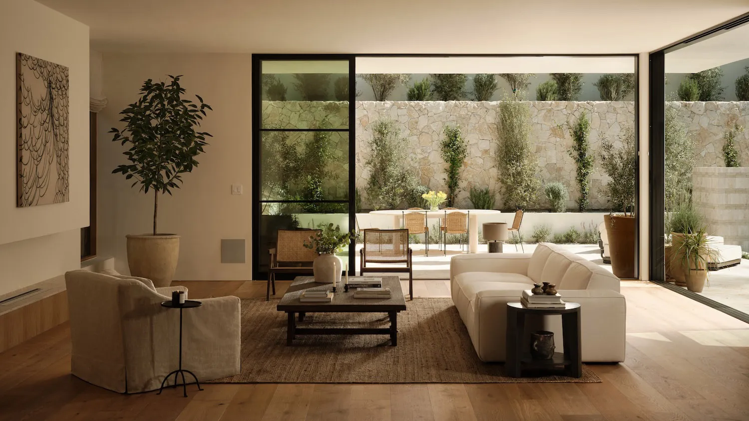 Modern neutral living room featuring a creamy sectional sofa, linen armchair, dark wood coffee table, large potted tree, and expansive glass doors opening to a sunlit courtyard with stone walls and outdoor dining area.