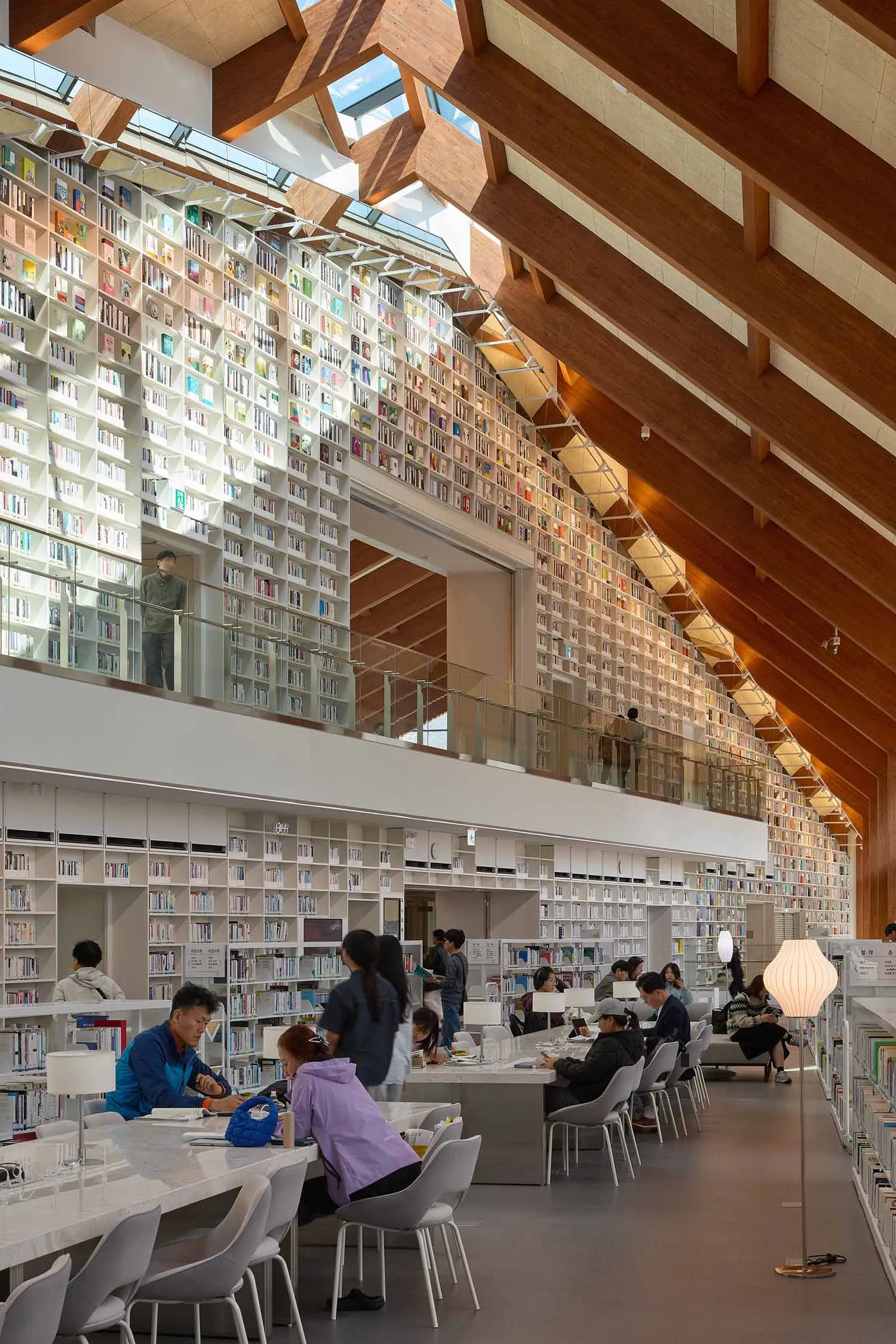 Interior view of a modern multi-level library with massive white bookshelves, warm wooden ceiling structure, students studying at long white tables, and people browsing on the upper balcony.