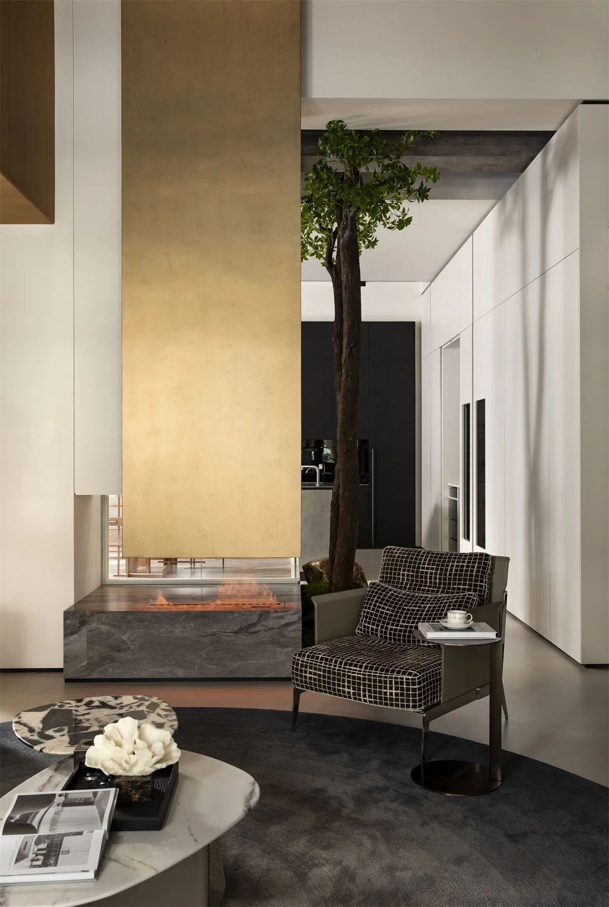 Modern minimalist living room featuring a tall indoor tree, a gold leaf and dark marble fireplace, a checkered armchair, and circular marble coffee tables on a dark rug.