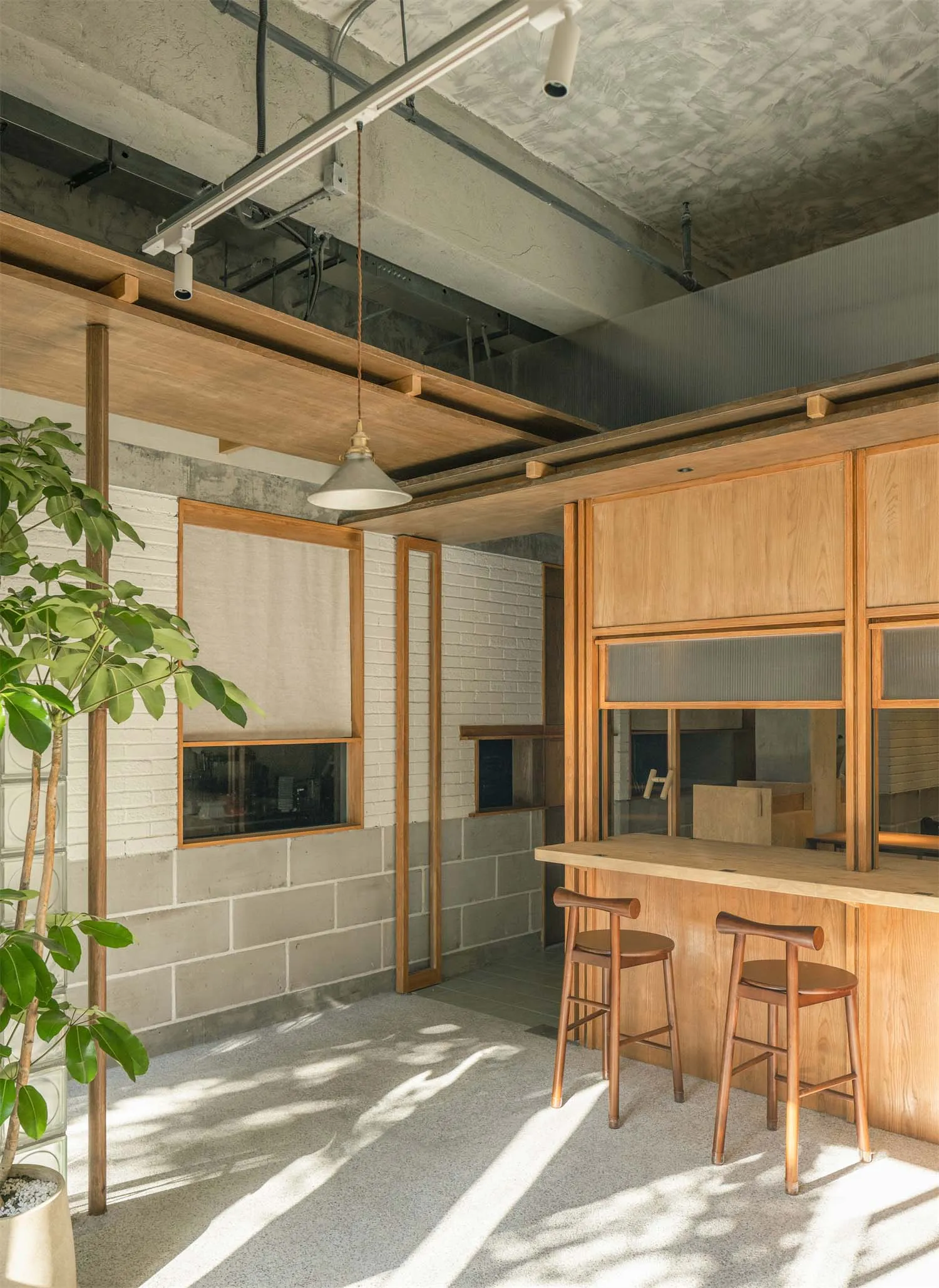 Modern industrial-style cafe interior with a long oak wooden bar counter, two matching oak stools, exposed concrete ceiling and block walls, wooden beams, and a large potted plant in the foreground.