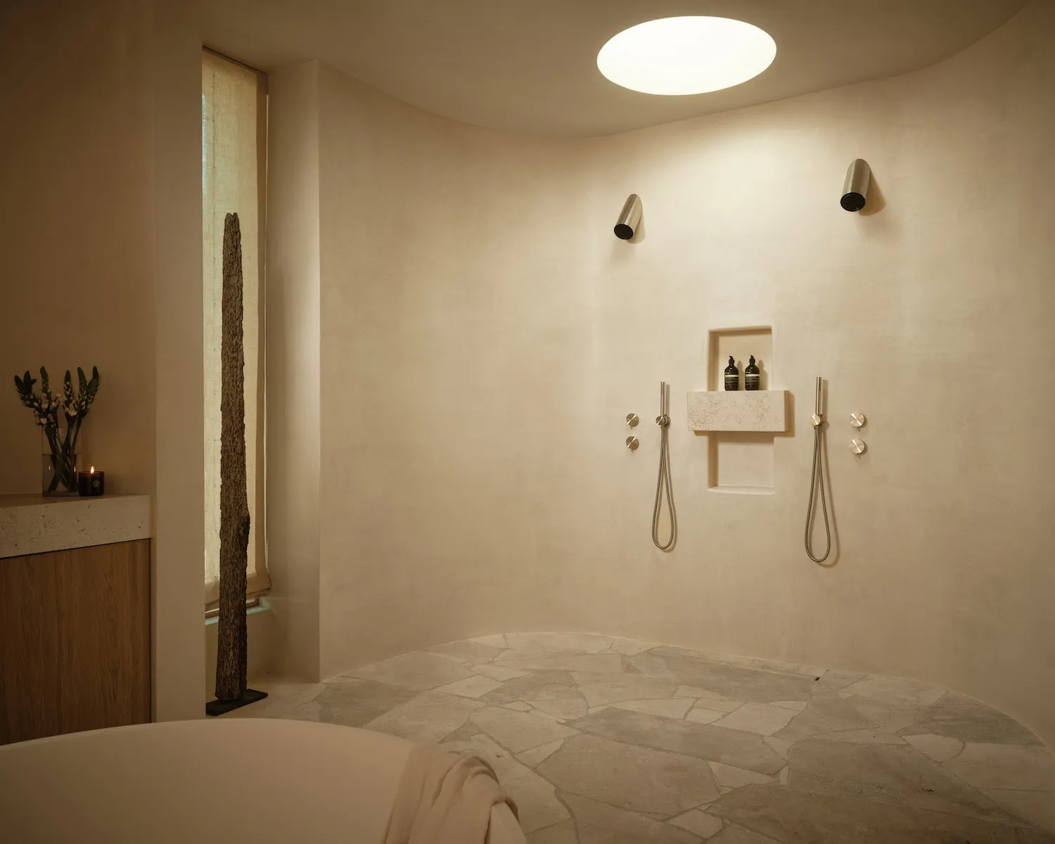 Modern minimalist curved bathroom with travertine stone floor, freestanding white tub, and integrated shower niche with brushed metal fixtures.