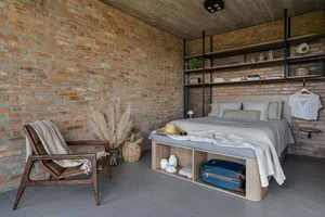 A modern, minimalist bedroom featuring brick and concrete walls, a wooden bed frame, and a leather chair.