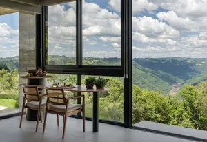 A modern dining room with a table, chairs, and a view of lush green hills through a large glass window.