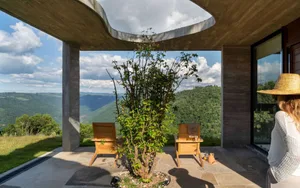 An outdoor area with concrete flooring and a tree, featuring two wooden chairs and a scenic mountain view.