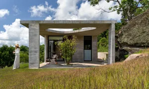 A modern residential structure made of concrete, featuring a patio with a small tree and a person wearing a hat and sunglasse