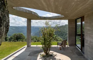 A modern residential interior featuring a concrete ceiling and floor, with a tree and chair on the patio.