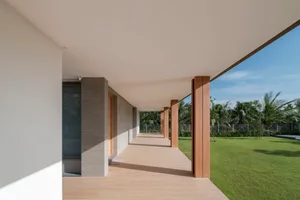 A modern architectural walkway with wooden pillars and concrete walls, surrounded by greenery and blue sky.