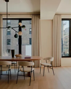 A modern dining room featuring a round oak table surrounded by upholstered chairs, large windows with curtains, and a chandel