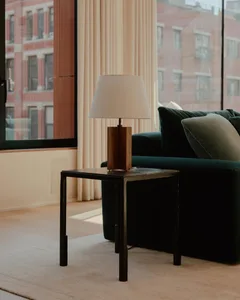 A modern minimalist living room featuring a wooden side table with a white lamp shade, a green velvet sofa, and a large windo