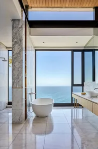 A modern marble bathroom with a view of the ocean features an oval-shaped standalone bathtub, a sink vanity, and large window