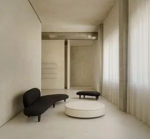 A minimalist living room featuring a black curved sofa, a round white ottoman, and a concrete floor.