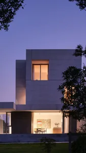 A modern, residential building with concrete exterior at dusk, featuring illuminated windows and a serene garden.