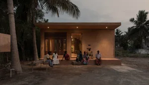 People sitting in front of a small clay home with a thatched roof in a tropical setting.