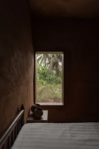 A minimalist bedroom with concrete walls, a bed, and a window overlooking greenery.