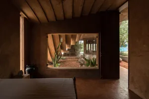 A modern bedroom featuring textured clay walls, concrete floors, and a window overlooking a courtyard with plants.