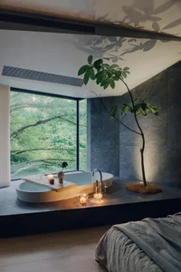 A modern minimalist bedroom featuring a stone bathtub and a potted plant, with a large window overlooking greenery.