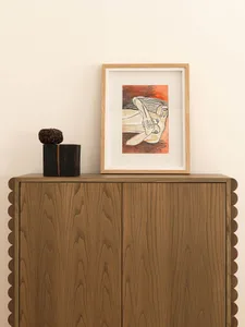 A modern Scandinavian-style scroller cabinet made from wood, topped with a framed piece of decorative art depicting a human f