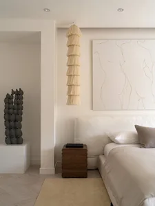 A minimalist bedroom with soft, neutral tones featuring a white bed, wooden nightstand, and fabric art hanging above the bed.