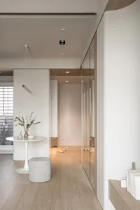 A minimalist interior design featuring a wooden floor with a round table, stool, and floral arrangement.