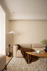 A cozy, minimalist living room featuring a beige sofa with a brown throw blanket, a wooden coffee table, a floor lamp, and a 