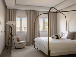 Modern bedroom featuring a fabric and metal bed frame, large windows with beige curtains, and a white upholstered chair with