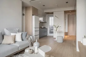 A minimalist living room with a grey sofa, wooden flooring, and a kitchen area in the background.