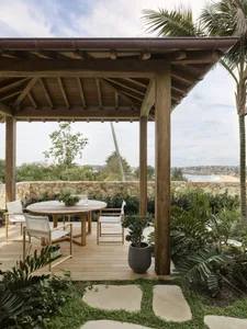 A wooden outdoor patio with a table and chairs under a wooden canopy, surrounded by greenery and overlooking a scenic landsca