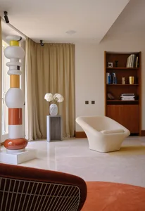 A modern interior with a white sculptural piece, a white armchair, and a vase of white flowers on a concrete pedestal.