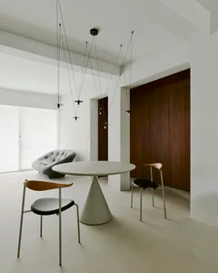 A modern dining room featuring a round, white, cone-shaped table with minimalist black chairs, and a gray tufted armchair in 