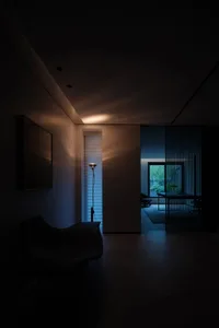 Modern, minimalist hallway with concrete walls and floor, featuring a single floor lamp casting a warm glow.