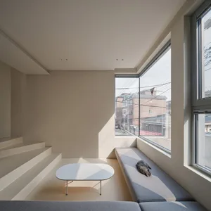 A modern minimalist living room with a grey couch, a white coffee table, and a window with a cat resting on the sill.