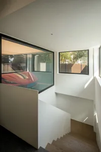 Modern residential staircase with white concrete walls and floor, featuring a large window revealing a sleek red sports car p
