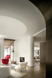 A modern living room with a white fur-covered chair, a round glass table, and a fireplace. The space features clean lines, mi
