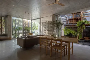 A modern living room featuring concrete walls and floor, with plants, a wooden table, and a brown sofa.