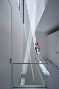 A woman stands on a glass wall railing in a modern, white interior space.