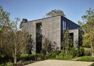 A modern, metal-clad house with large windows and a gravel driveway surrounded by greenery and young trees.