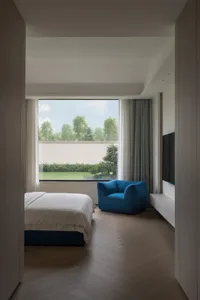 A modern minimalist bedroom with a wooden floor, a white bed, and a blue armchair, featuring a large window with a view of tr