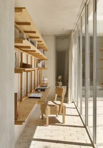 A minimalist home office featuring wooden furniture, shelves, and a desk with books and a lamp.