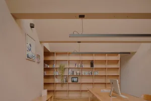 A minimalist home office with wooden shelves filled with books and plants, featuring a desk with a computer and keyboard.