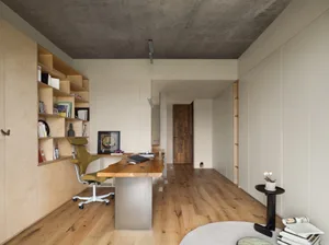 A modern minimalist office space featuring a wooden desk and shelving unit against a concrete and beige wall.