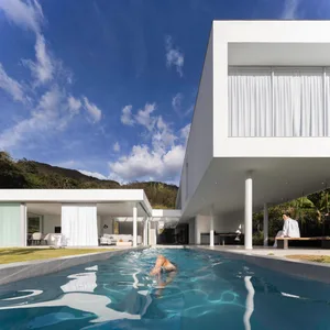 A modern minimalist house with white exterior, large windows, and a Swimming Pool. A person is swimming in the pool, while an