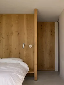 A minimalist bedroom featuring wooden paneled walls, a simple bed with white bedding, and a wall-mounted lamp.