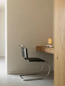 A minimalist black leather chair with chrome frame on a hardwood floor, next to a wooden desk with an open book and a small l
