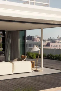 A minimalist living room with a white sofa, a white floor lamp with a curved base, and a wooden deck overlooking a cityscape.