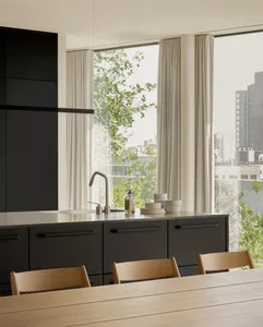 A modern kitchen with black metal cabinets, a wooden dining table, and large windows overlooking greenery and urban buildings