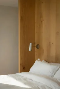 A minimalist bedroom with white bedding, wooden walls, and a wall-mounted lamp.