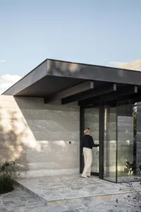 A woman standing at the entrance of a modern house with concrete walls, stone floor, and glass door.