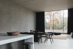 A modern living room with concrete walls and floor, featuring a large window with black curtains, a black wooden dining table