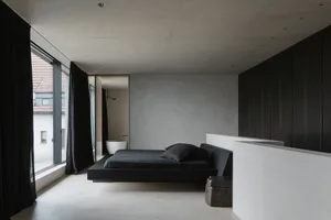 A modern, minimalist bedroom with a concrete floor and walls, featuring a low-profile bed and large windows.